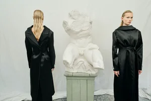 Two models in long black coats flanking a classical white torso sculpture against a plain backdrop.