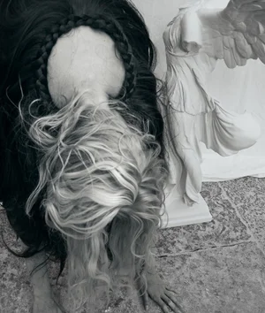 Model with long dark and blonde hair bent over next to a white winged statue on a textured floor.