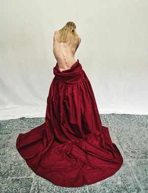 Model with long blonde hair seen from behind, wearing a voluminous red gown draped low on the hips against a white backdrop.