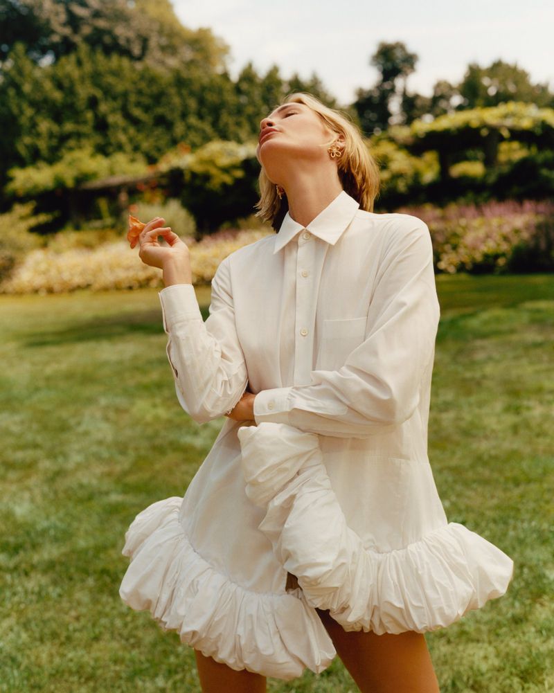 Fashion shot of a blonde woman in a white dress standing in a grassy field with trees. Outdoor fashion photography in nature.