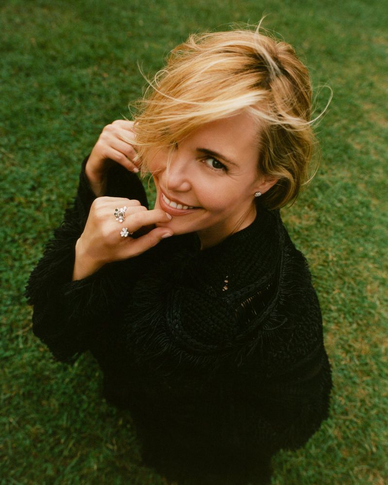 Smiling woman with short blonde hair, wearing a black top and rings, stands in a green grassy field, captured from a high angle.