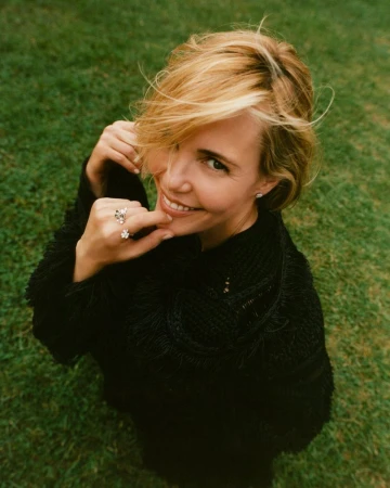 Smiling woman with short blonde hair, wearing a black top and rings, stands in a green grassy field, captured from a high angle.