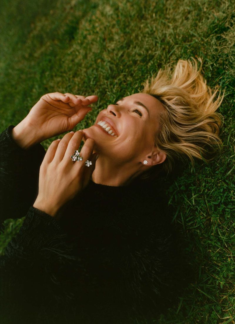 Leslie Bibb lies on green grass smiling with hands raised near her face, wearing a black textured knit top and sparkling rings.