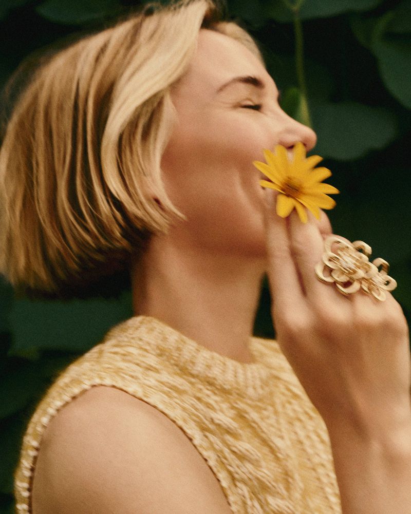 Close-up of a blonde adult female smiling and holding a yellow flower to her nose, wearing a yellow sweater and a large gold ring.