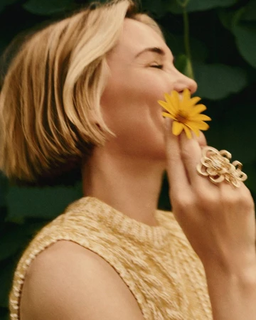 Close-up of a blonde adult female smiling and holding a yellow flower to her nose, wearing a yellow sweater and a large gold ring.
