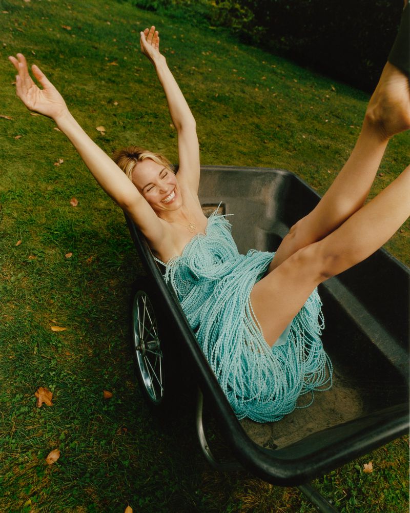 A young adult woman with blonde hair in a light blue dress lies in a metal wheelbarrow on a green grass lawn with arms outstretched.