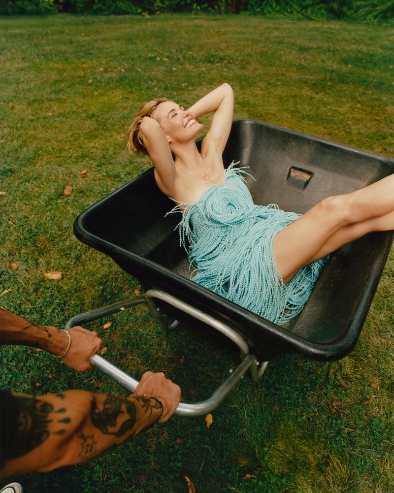 Woman in a blue fringed dress reclines in a wheelbarrow being pushed on a lawn with trees in the background on a sunny day.