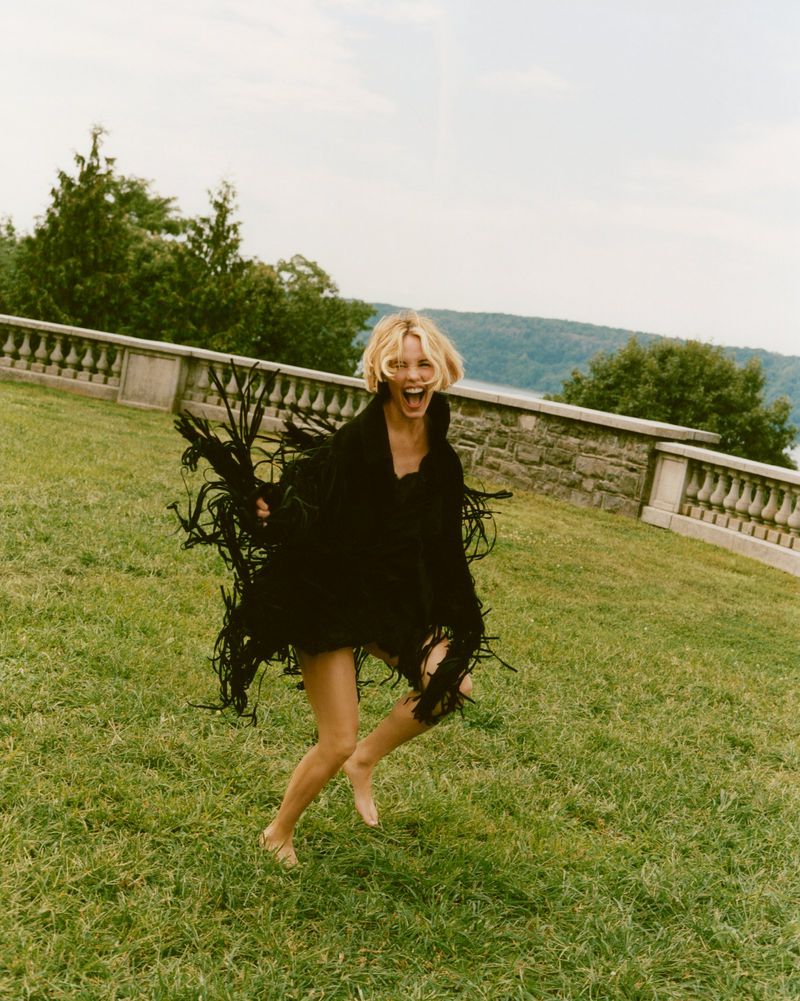 Woman with blonde hair jumps joyfully on a grassy lawn wearing a black fringed shawl, with stone architecture and trees in the background.