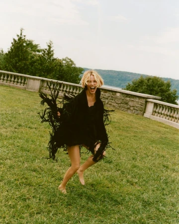 Woman with blonde hair jumps joyfully on a grassy lawn wearing a black fringed shawl, with stone architecture and trees in the background.