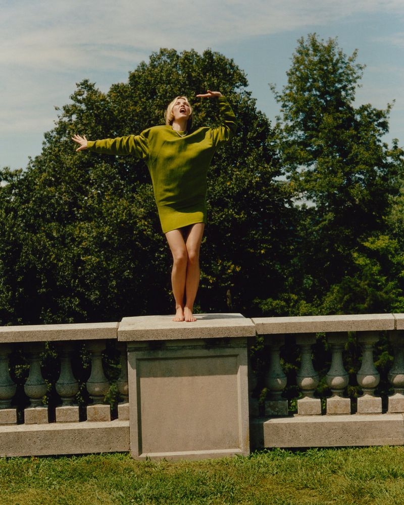 Woman in green oversized sweater stands on stone wall with arms outstretched, photographed outside with trees and blue sky in the background.