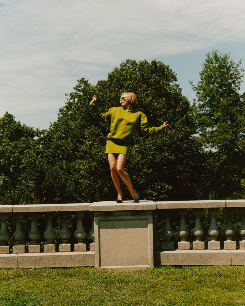 Woman in a green skirt and sweater standing on a stone wall outdoors. Green trees and blue sky create a vibrant, natural setting.
