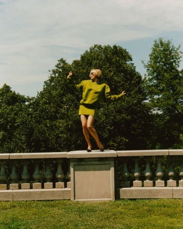 Woman in a green skirt and sweater standing on a stone wall outdoors. Green trees and blue sky create a vibrant, natural setting.