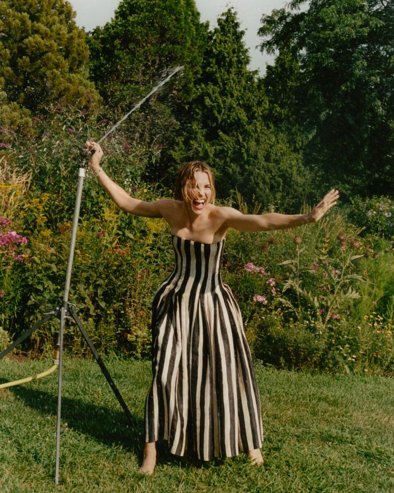 A woman in a striped dress stands in a garden, arms outstretched, holding a hose that sprays water, with lush greenery surrounding her.