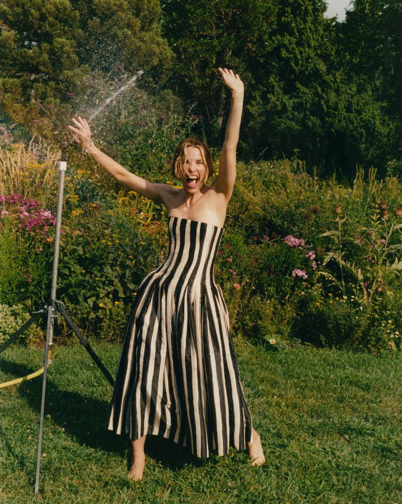 Adult woman in a black-and-white striped strapless dress stands in a grassy yard with a garden sprinkler and trees behind her.