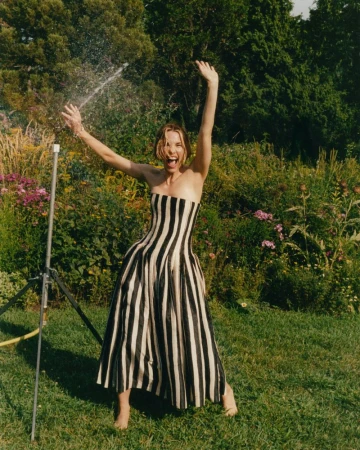 Adult woman in a black-and-white striped strapless dress stands in a grassy yard with a garden sprinkler and trees behind her.
