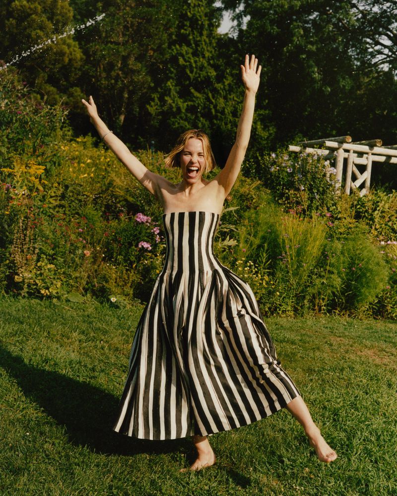 Woman in a black-and-white striped dress jumps on green grass in a garden setting with greenery, plants, and trees behind her.