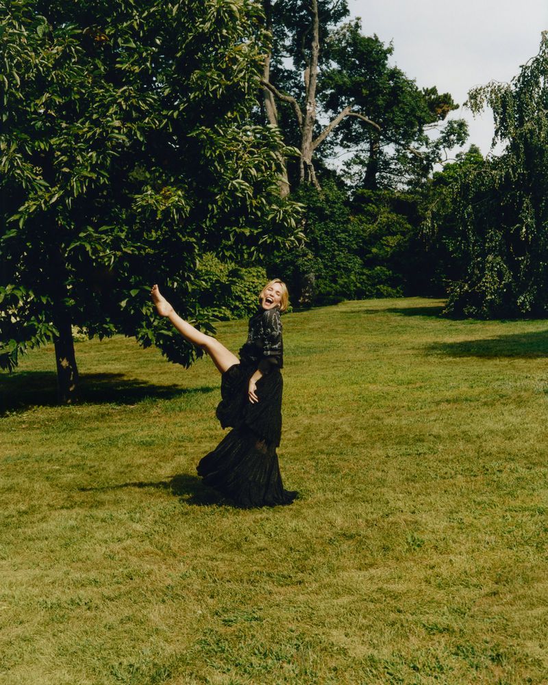 Young woman in black dress with one leg raised in a grassy, green park under trees with visible sky. Fashion editorial photoshoot.