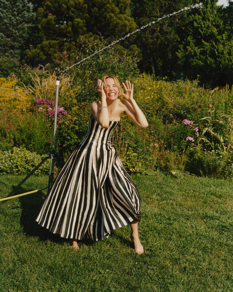 Adult woman in a striped dress standing in a yard with sprinkler, smiling with hands on face. Green lawn and foliage background.