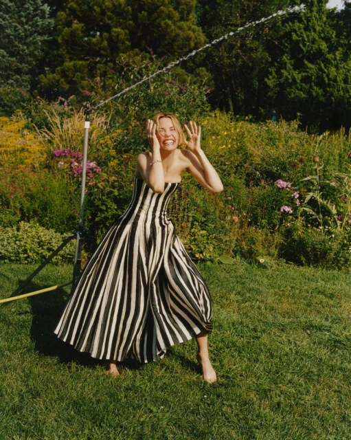 Adult woman in a striped dress standing in a yard with sprinkler, smiling with hands on face. Green lawn and foliage background.