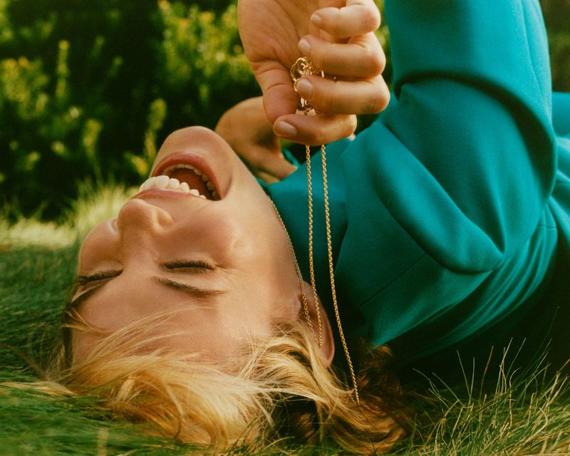 Leslie Bibb laughing while lying on green grass wearing a teal jacket holding a gold necklace for Marie Claire Australia