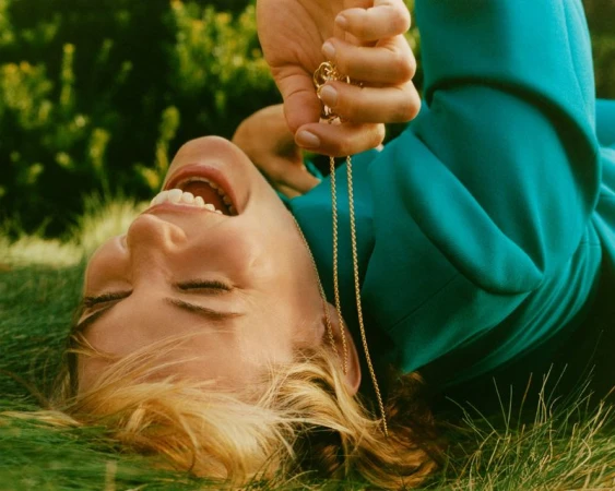Leslie Bibb laughing while lying on green grass wearing a teal jacket holding a gold necklace for Marie Claire Australia