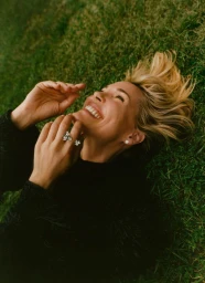 Leslie Bibb lies on green grass smiling with hands raised near her face, wearing a black textured knit top and sparkling rings.