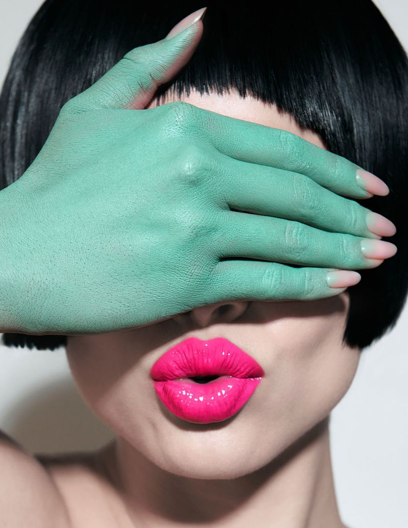 Close-up portrait of a young adult woman with a short black bob, pink lips, and a mint green-painted hand covering her eyes.