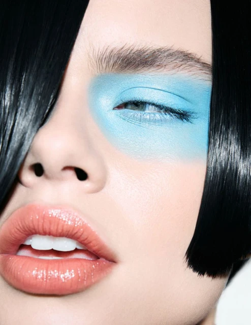 Close-up beauty shot of a woman with black hair and blue eyeshadow, showcasing glossy lips and fair skin against a white background.