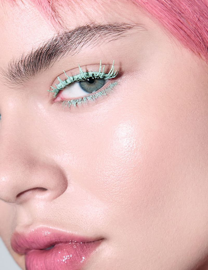 Close-up of a woman's face showcasing creative makeup: vibrant pink hair, flawless skin, glossy pink lips, and striking green mascara.