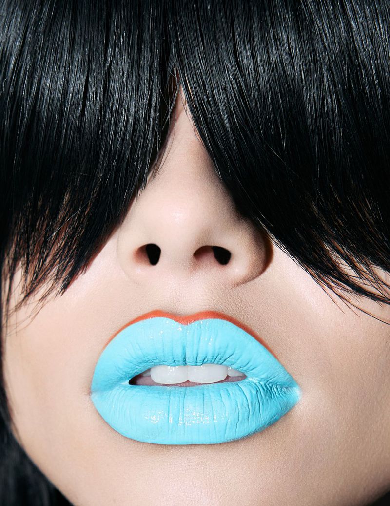 Close-up of a person's face with bright blue lipstick and black hair obscuring the forehead. Beauty shot emphasizing facial features.