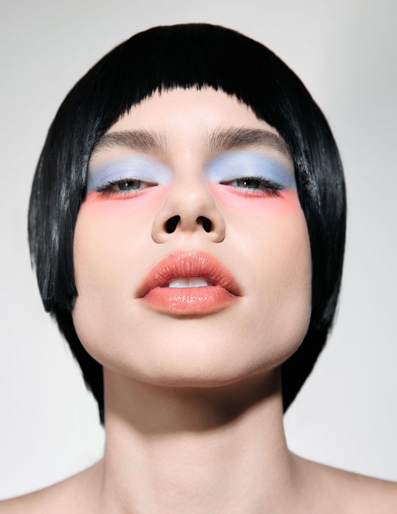 Headshot of a woman with a blunt black bob, blue eyeshadow, and pink blush, looking directly at the camera against a white background.