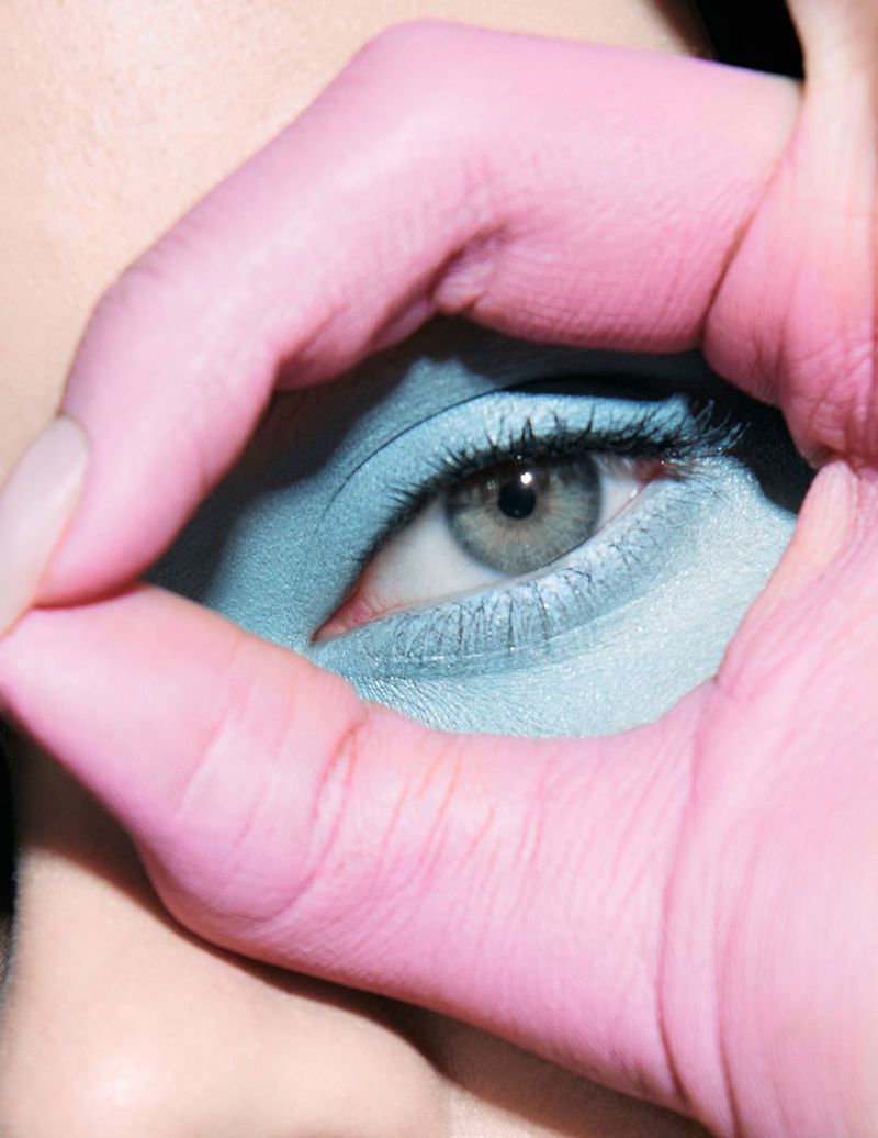 Close-up of a woman's face with blue eyeshadow, framing her gray eye with a pink-toned hand in a circular gesture, minimalist beauty.