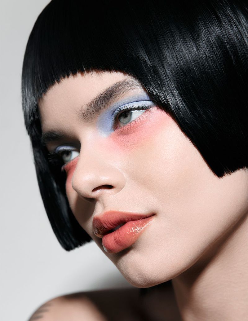 Woman with short black hair and stylized pastel makeup posing in a beauty shot with a bright neutral background.