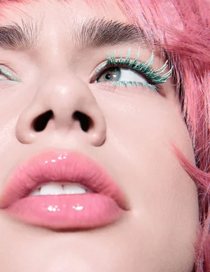 Extreme close-up beauty portrait of a person with vibrant pink hair, mint green eyelashes, and glossy pink lips.