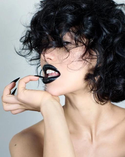 Close-up of a young adult woman with black curly hair and dark lipstick, holding a thin black object near her mouth against a light background.