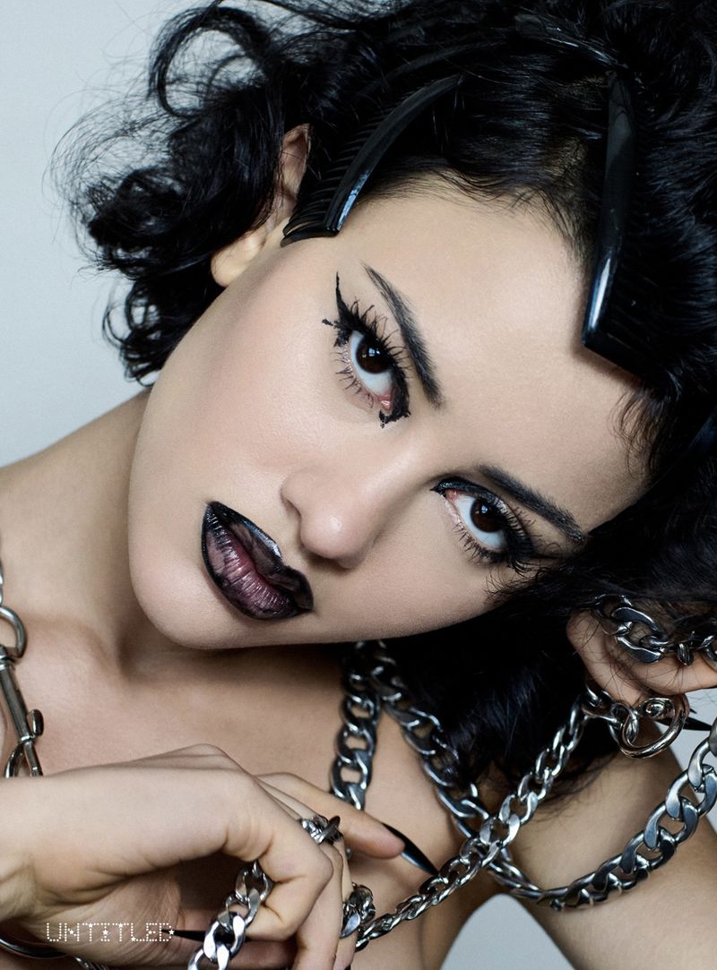 Close-up studio shot of a woman with dark curly hair and dramatic makeup, wearing a heavy silver chain necklace. Dark lip color.