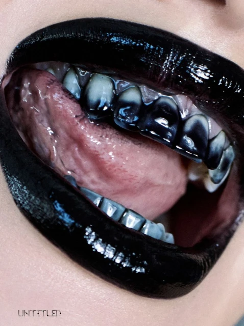 Macro photograph of a woman's mouth featuring black lipstick, exposed teeth with blue and black accents, and a visible tongue.