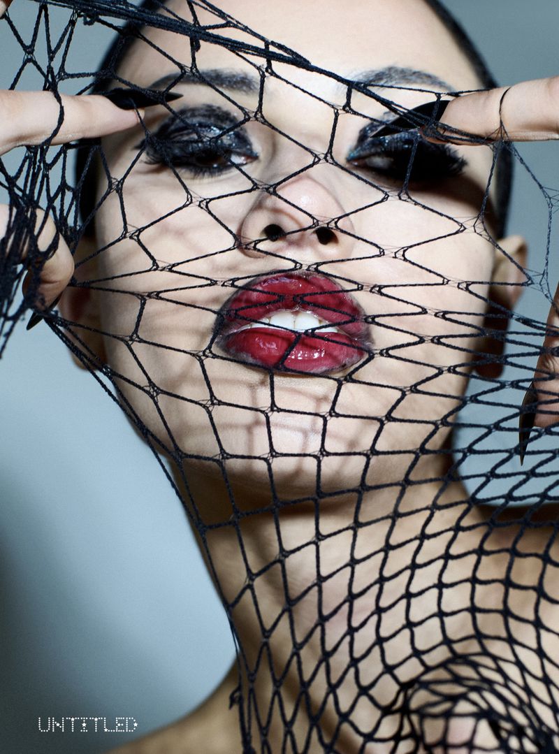 Close-up portrait of a woman with dramatic eye makeup and bold red lips posing behind a black fishnet veil, creating a striking fashion image.