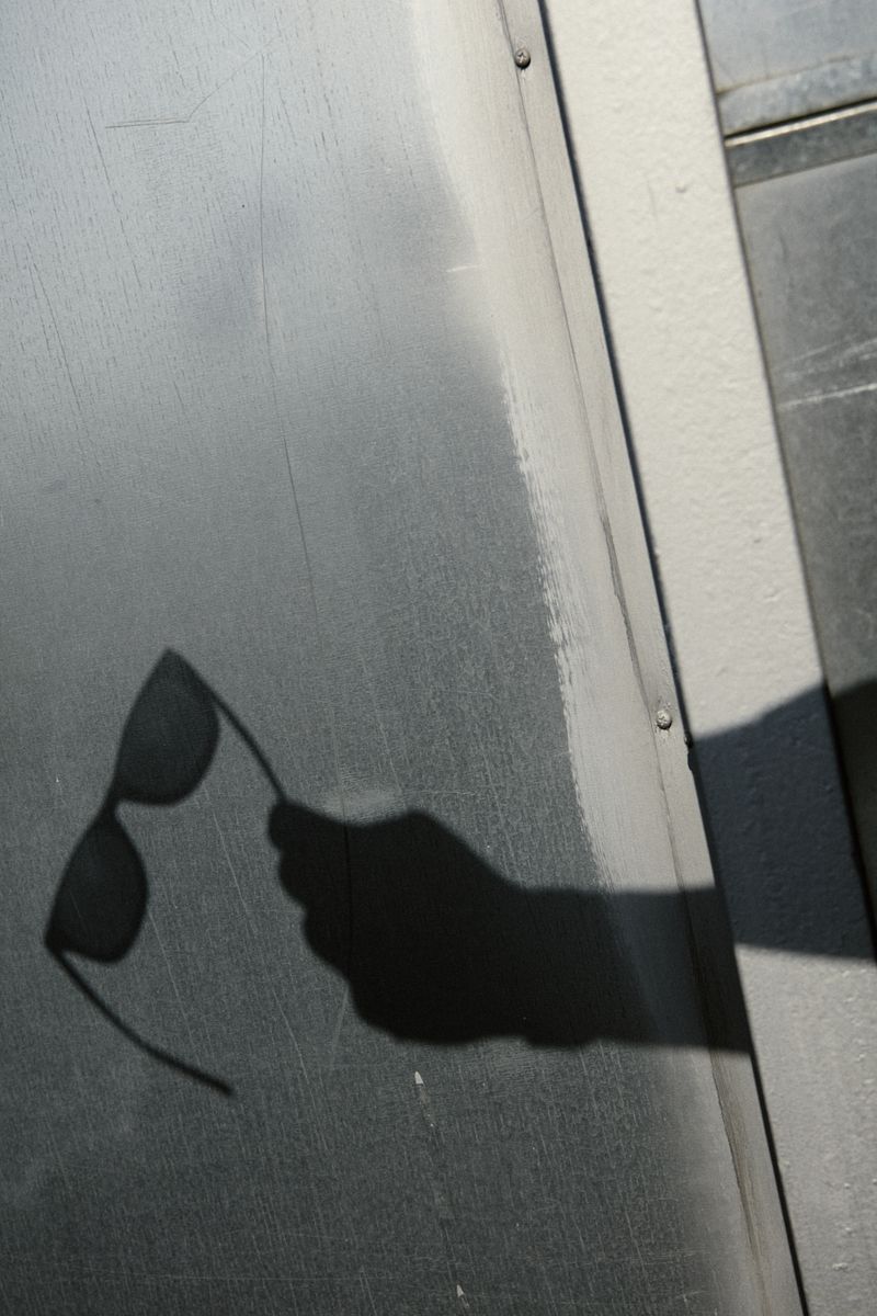 Shadow of a hand holding sunglasses is cast onto a gray metal surface, creating a stark contrast in light and shadow.