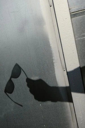 Shadow of a hand holding sunglasses is cast onto a gray metal surface, creating a stark contrast in light and shadow.