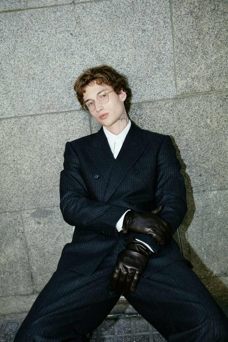 Man in a dark pinstripe double-breasted suit with black gloves poses against a grey concrete wall in an outdoor setting.
