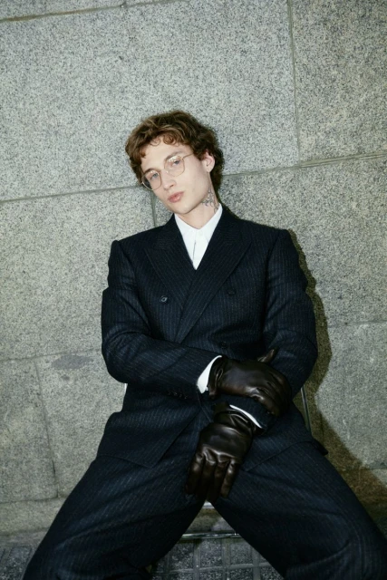 Man in a dark pinstripe double-breasted suit with black gloves poses against a grey concrete wall in an outdoor setting.