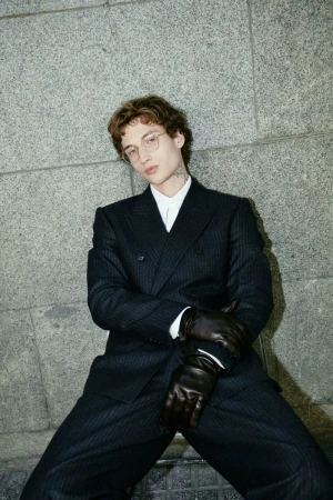 Man in a dark pinstripe double-breasted suit with black gloves poses against a grey concrete wall in an outdoor setting.