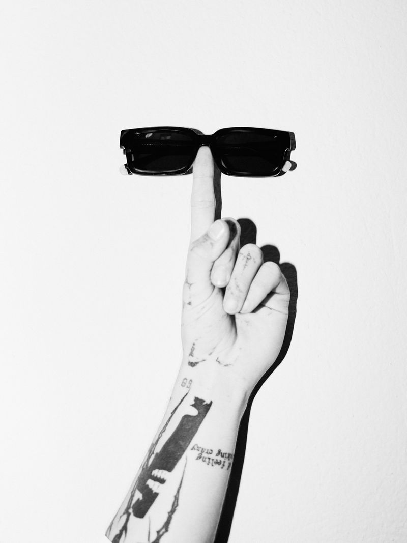 Sunglasses balanced on a finger in a black-and-white studio shot, showcasing fashion eyewear with a tattoo on the arm, and a clean aesthetic.