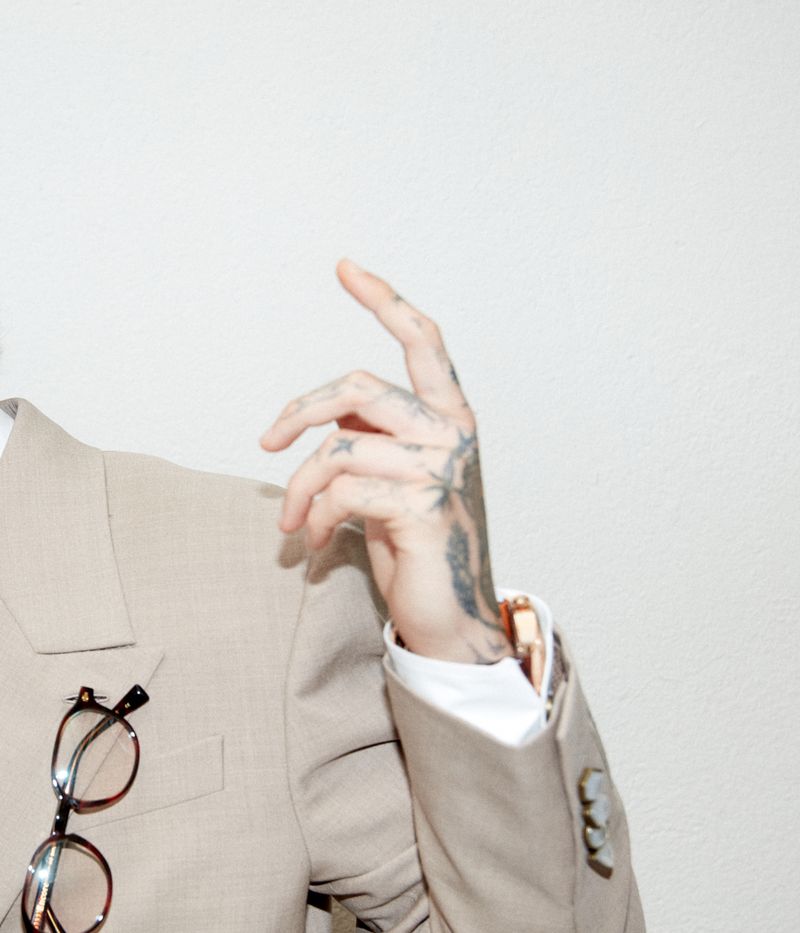 Close-up of a tattooed hand, a beige blazer with eyeglasses, and a gold watch against a white background.