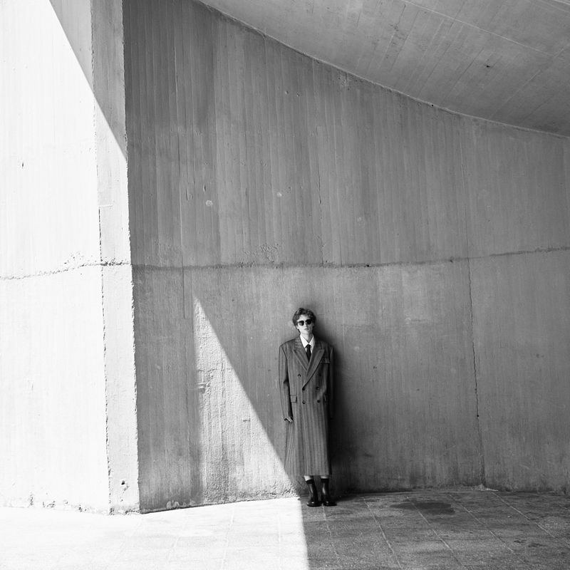 Black and white portrait of a slender adult in a coat and sunglasses, posing in front of a concrete wall in an urban underpass.