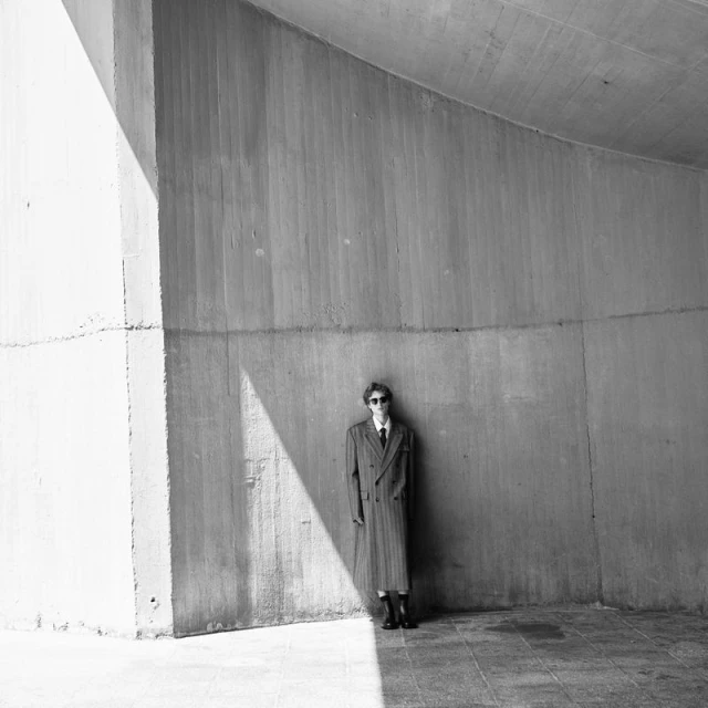 Black and white portrait of a slender adult in a coat and sunglasses, posing in front of a concrete wall in an urban underpass.