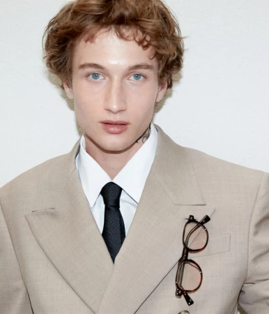 Man in a tan suit with a black tie poses indoors against a white background. He has light skin, reddish hair, and light blue eyes.