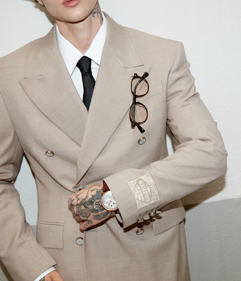 Cropped shot of a man wearing a double-breasted beige suit jacket with a black tie, eyeglasses on the lapel, and visible hand tattoos.