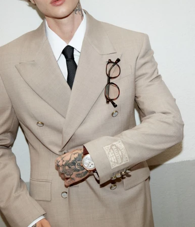Cropped shot of a man wearing a double-breasted beige suit jacket with a black tie, eyeglasses on the lapel, and visible hand tattoos.